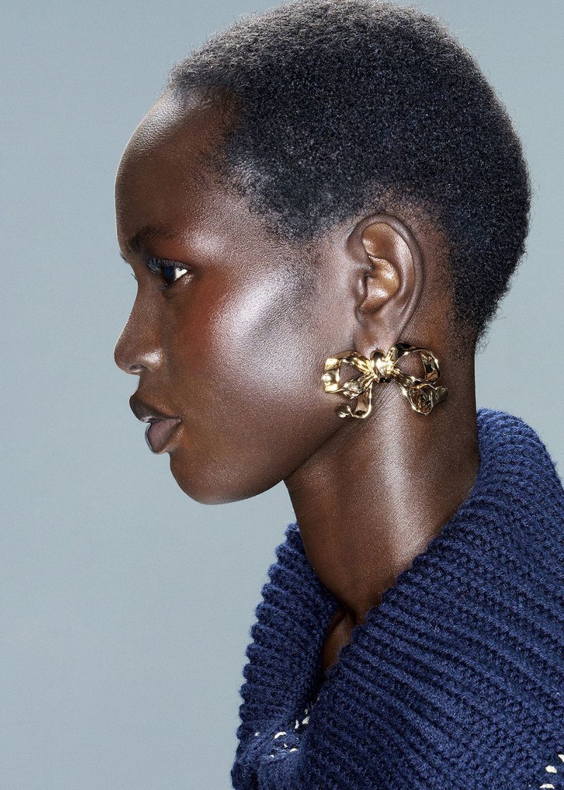 Close-up profile of an adult woman with dark skin, wearing a blue sweater and decorative gold bow earrings, on a grey background.