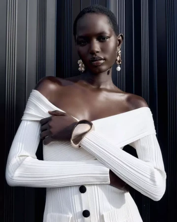 Model Nyaduola Gabriel wearing an off-the-shoulder white ribbed knit dress and gold cuff against a dark fluted backdrop.