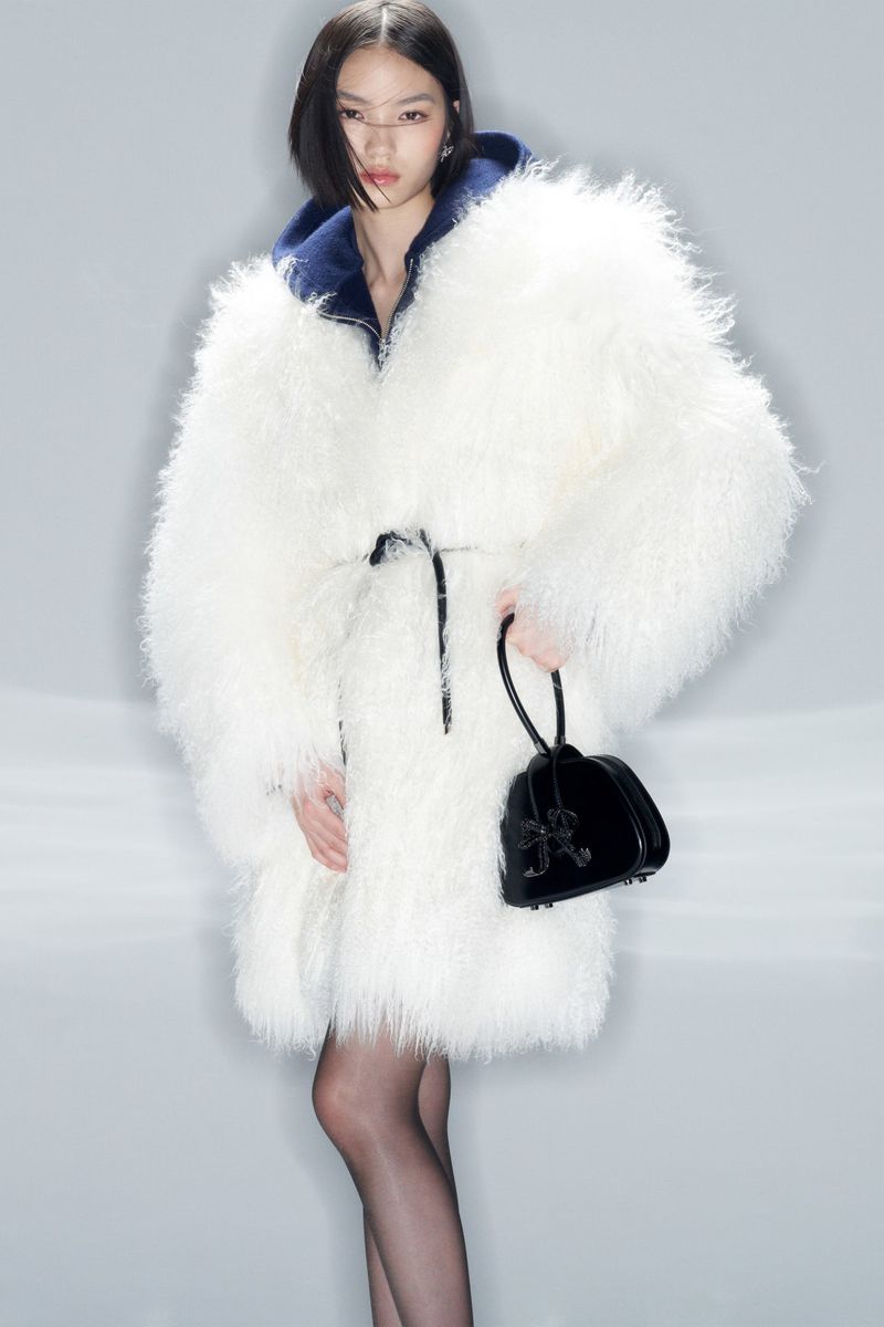 Fashion shot of a stylish Asian woman wearing a large white fur coat, black sheer tights, and carrying a shiny black purse with a bow detail.