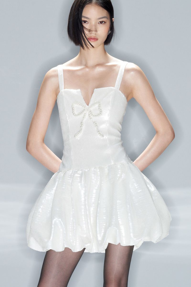 Young woman poses indoors wearing a white, bow-adorned mini dress and tights in a studio setting, showcasing a youthful and elegant fashion style.