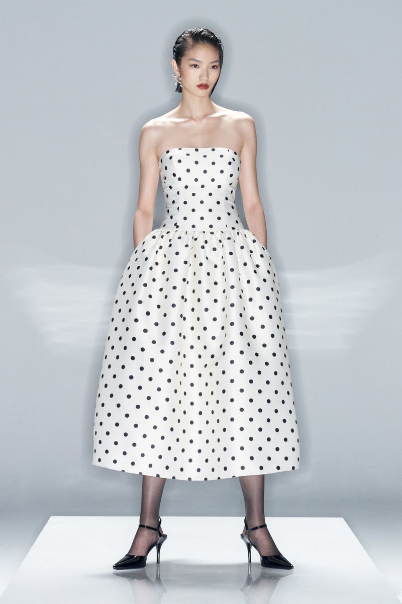 An Asian female fashion model showcases a strapless, tea-length, polka-dot dress with black tights and heels in a photo studio setting.