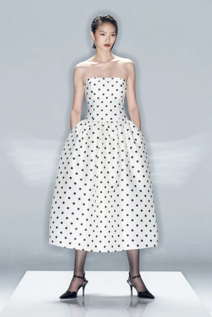 An Asian female fashion model showcases a strapless, tea-length, polka-dot dress with black tights and heels in a photo studio setting.