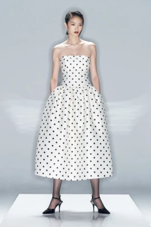 An Asian female fashion model showcases a strapless, tea-length, polka-dot dress with black tights and heels in a photo studio setting.