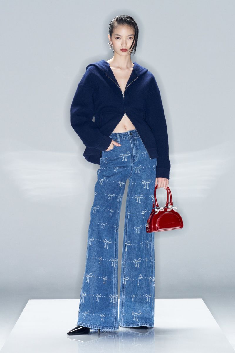 Asian woman wearing flared jeans with bows and a blue hooded top, holding a red purse, stands in a studio with a white backdrop.