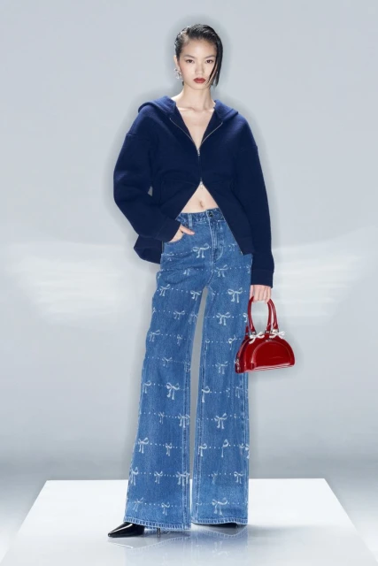 Asian woman wearing flared jeans with bows and a blue hooded top, holding a red purse, stands in a studio with a white backdrop.