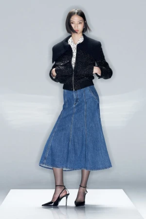 Full-shot of a woman in a black jacket, polka-dot shirt, and denim skirt, posing in black heels in a studio with a neutral background.
