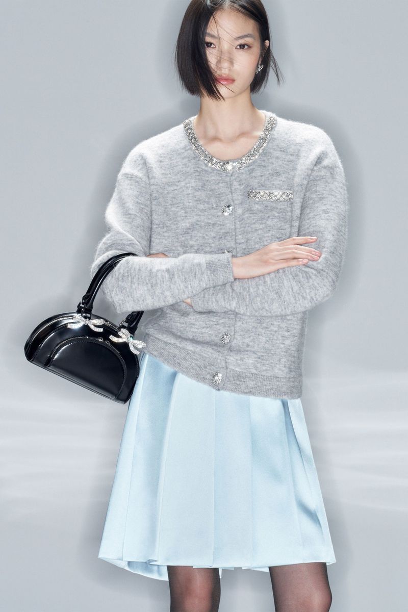 Portrait of an Asian woman in a gray cardigan, light blue skirt, and black purse posing in a studio with a neutral gray background.