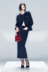 Model Huijia Chen in a navy skirt suit holding a red handbag against a seamless grey studio backdrop