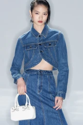 Model wearing a cropped blue denim jacket and matching skirt holding a small white handbag against a pale grey studio background.