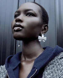 Close-up portrait of a model wearing sparkling bow earrings and a blue hoodie against a dark ribbed background.