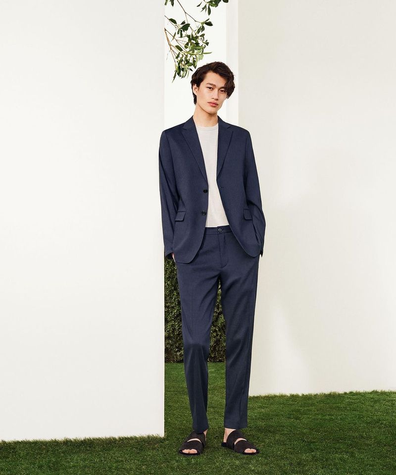 A young adult man in a dark blue suit and casual sandals stands in front of a white wall, complemented by green grass and foliage.