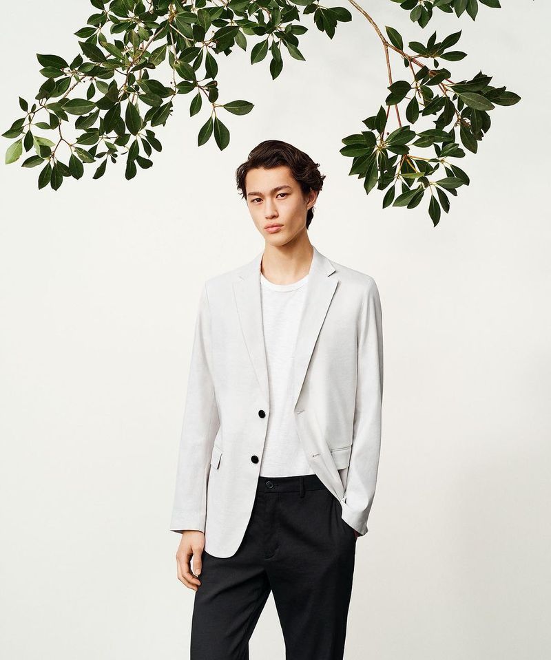 Portrait of a young adult male in a blazer and trousers, posing under a leafy branch against a white backdrop in a studio shot.