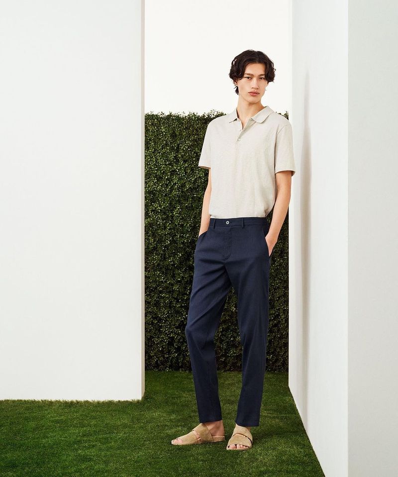 Man poses wearing casual clothing in an indoor photo studio with faux grass and hedge wall. He wears a beige polo shirt and navy trousers.