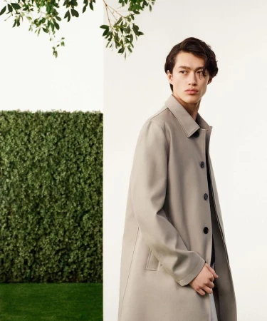A stylish Asian man in a trench coat poses against a hedge backdrop, showcasing fashion in a bright, outdoor setting.