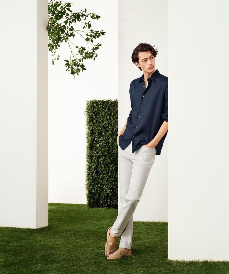 Man in a navy button-down shirt and white pants leaning against a white wall in an outdoor setting with green grass and foliage.