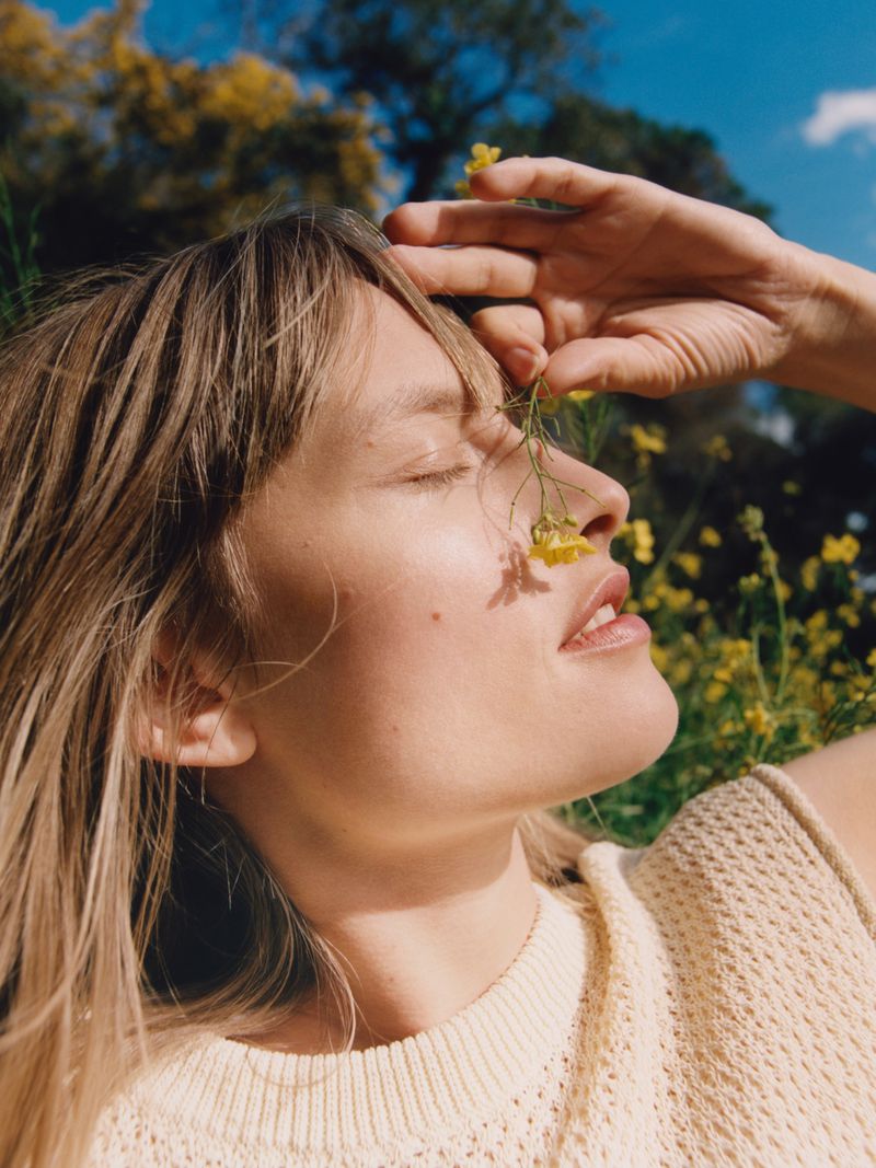 Woman with blonde hair poses in a field, enjoying the sunshine with a yellow flower resting against her nose and forehead.