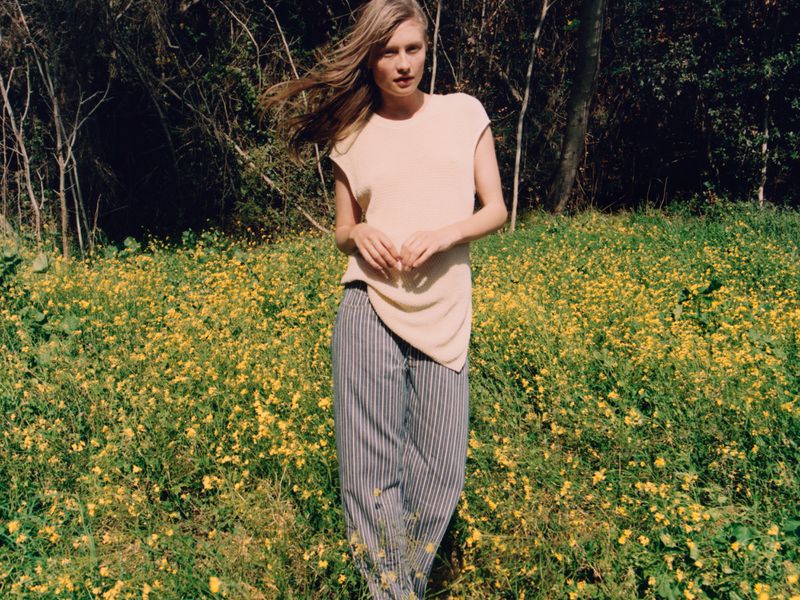 A blonde model stands in a field of yellow flowers wearing a light tan top and vertically striped gray and white pants in natural light.