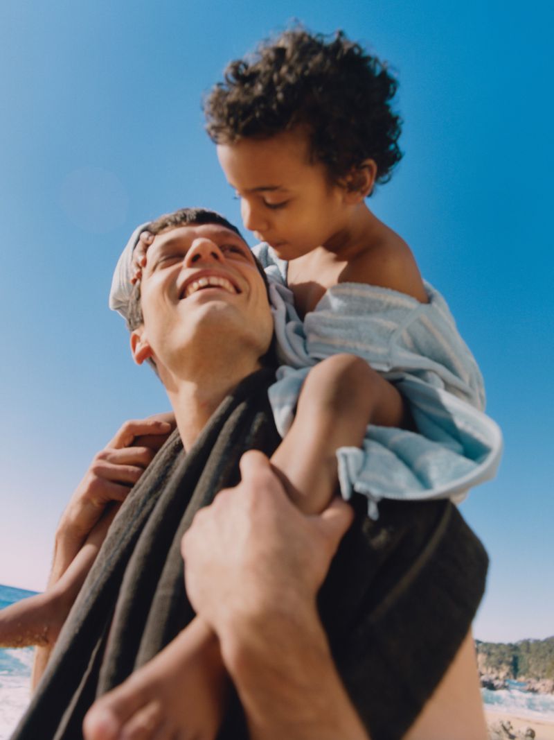 Man carrying child on shoulders at beach with blue sky. Father and daughter enjoying sunny day outdoors, creating a joyful moment.