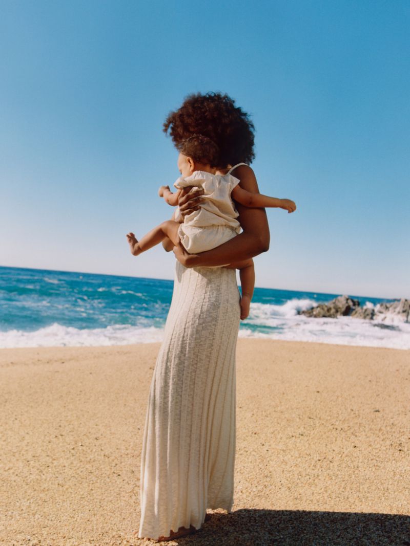 A mother holds her baby, backs to camera, on a sandy beach with the ocean and blue sky in the background on a sunny day.