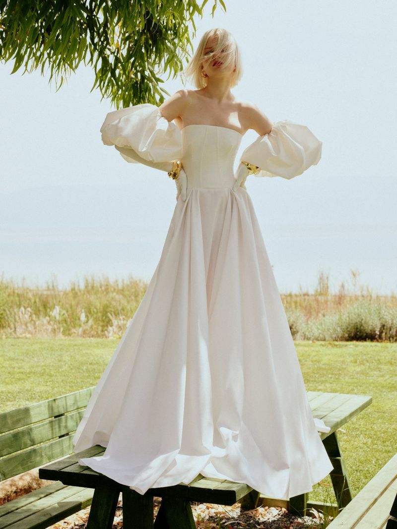 A woman in a white dress with large sleeves stands on a green picnic table in an outdoor grassy setting with trees overhead.