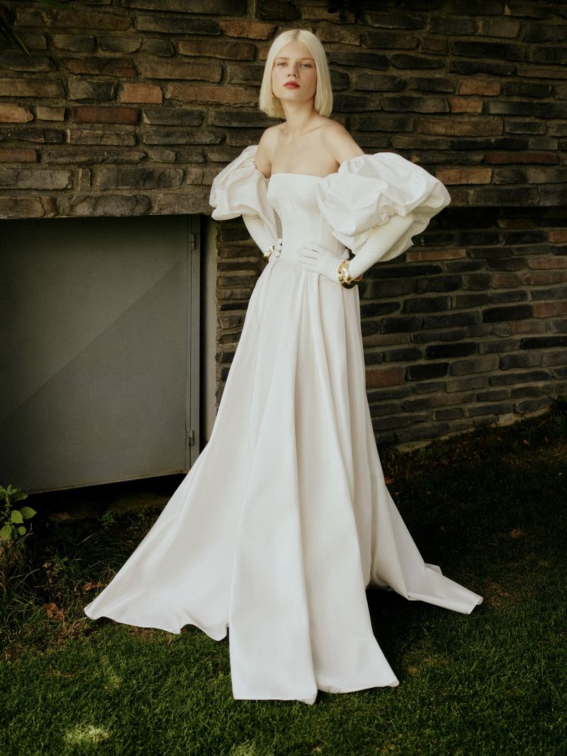 A blond woman poses in a white strapless gown with puff sleeves outside against a brick wall and green grass, showcasing fashion and elegance.