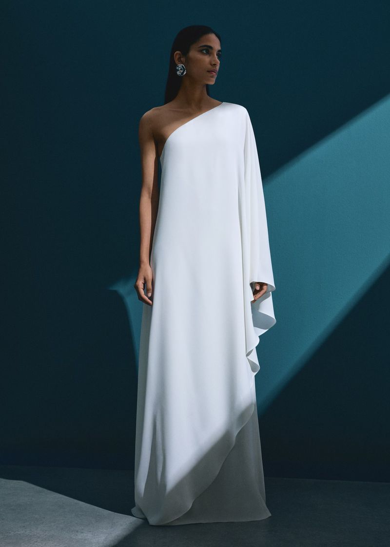 Woman in a flowing white, one-shoulder gown, posing in a studio with soft lighting creating a spotlight on her elegant attire.