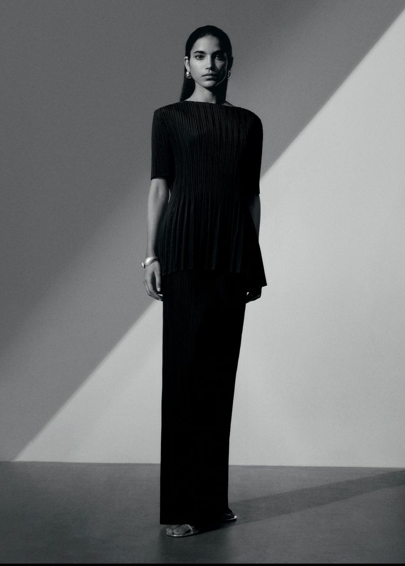 Fashion shoot featuring a South Asian female model in a dark, pleated top and long skirt against a gradient gray background with shadow.