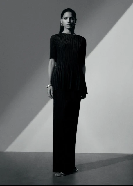 Fashion shoot featuring a South Asian female model in a dark, pleated top and long skirt against a gradient gray background with shadow.
