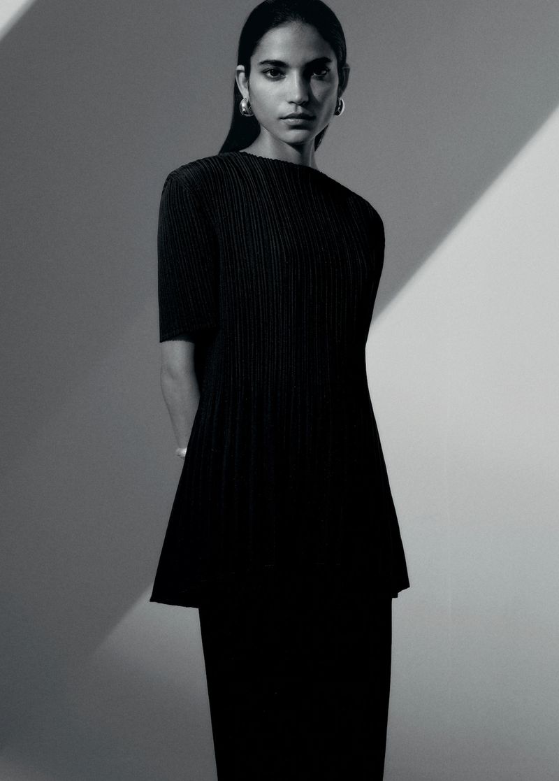 Studio portrait of a woman in a black pleated dress with shoulder-length hair, wearing hoop earrings against a grey backdrop.