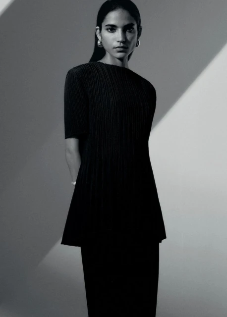 Studio portrait of a woman in a black pleated dress with shoulder-length hair, wearing hoop earrings against a grey backdrop.