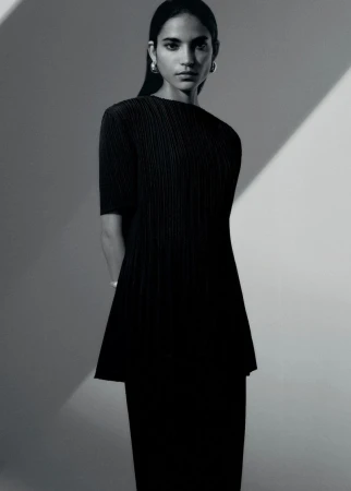 Studio portrait of a woman in a black pleated dress with shoulder-length hair, wearing hoop earrings against a grey backdrop.