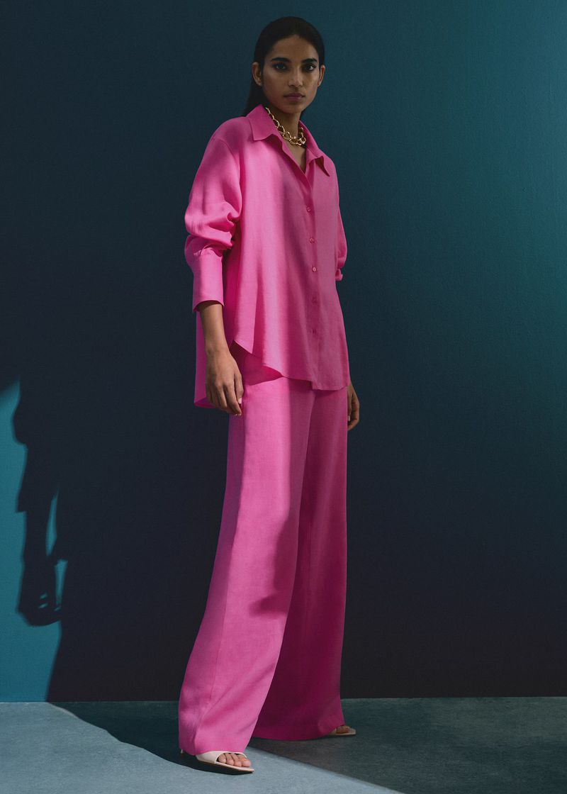 A woman poses in a hot pink long-sleeved button-down shirt and palazzo pants with bare feet against a dark teal background.