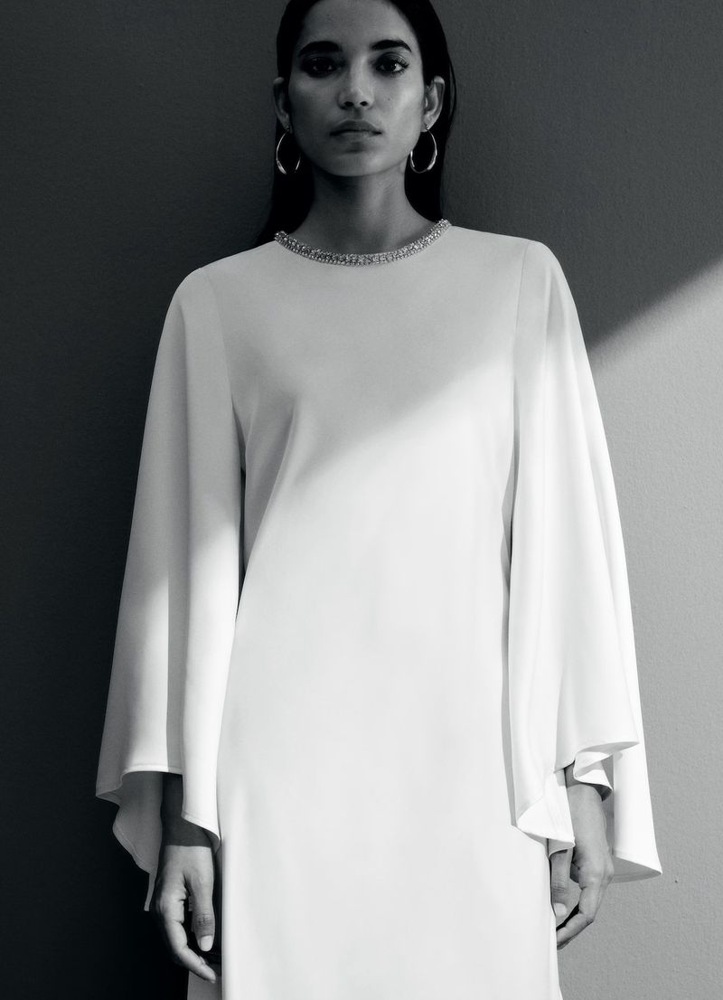 Model Amrit wearing a white cape-sleeve evening dress with an embellished neckline against a studio wall with sharp diagonal shadow.
