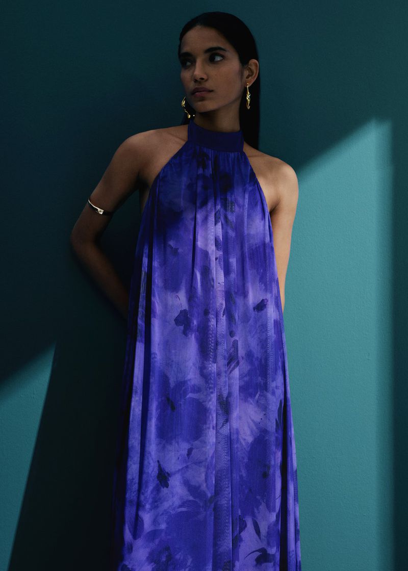 Woman in a halter dress with a floral pattern against a teal wall. She is wearing gold jewelry and has a neutral expression.