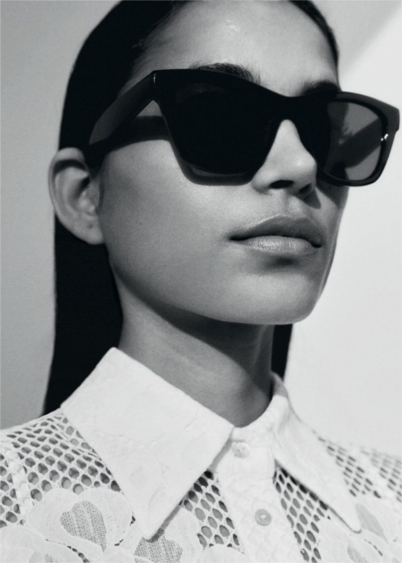 Close-up fashion shot of an adult female wearing sunglasses and a white lace collared shirt with a neutral facial expression in a photo studio.