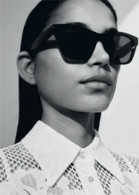 Close-up fashion shot of an adult female wearing sunglasses and a white lace collared shirt with a neutral facial expression in a photo studio.