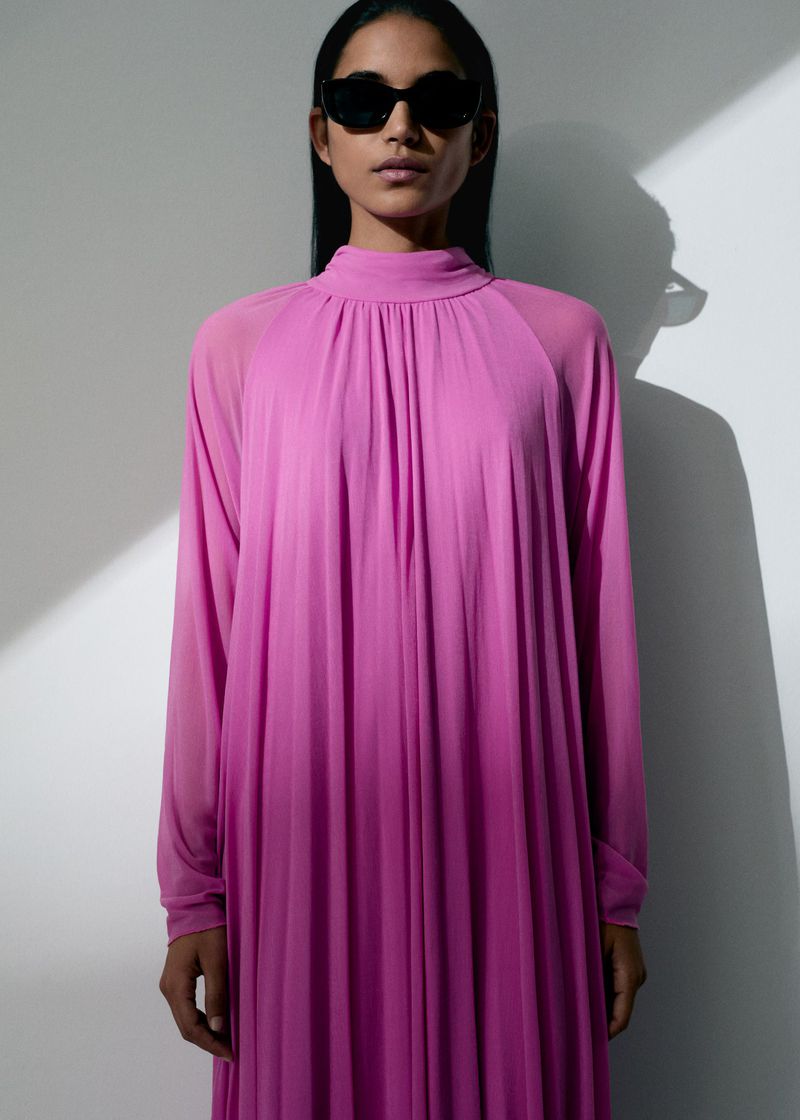 An adult woman in a long, pink dress with sunglasses stands indoors against a white wall in a fashion or beauty editorial shot.