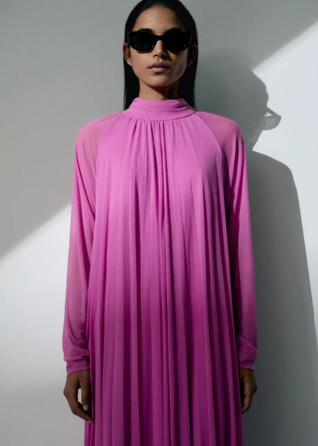 An adult woman in a long, pink dress with sunglasses stands indoors against a white wall in a fashion or beauty editorial shot.