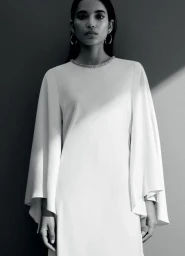 Model Amrit wearing a white cape-sleeve evening dress with an embellished neckline against a studio wall with sharp diagonal shadow.