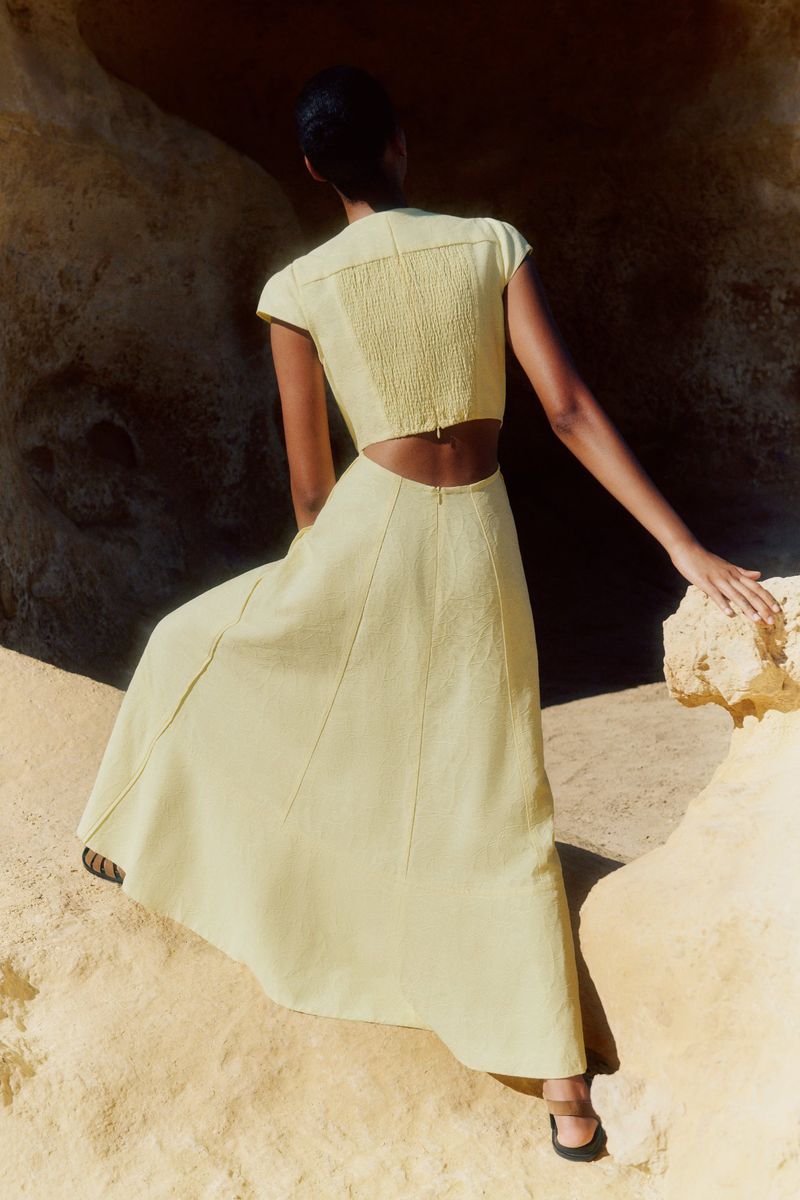 Woman in yellow dress stands on rocks, back to camera, minimal style and earthy tones, fashion editorial on location, outdoor setting.