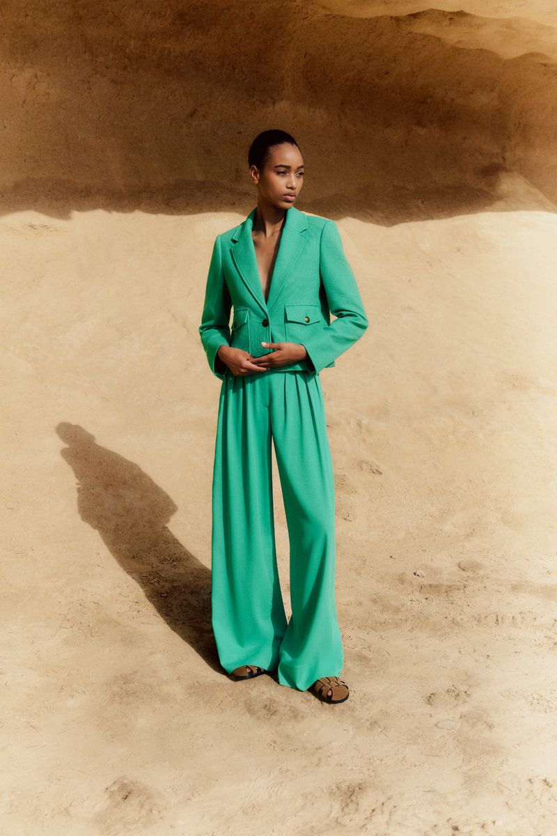 An adult female model poses in a bright turquoise-green pantsuit outdoors against a sandy-colored wall with natural lighting.