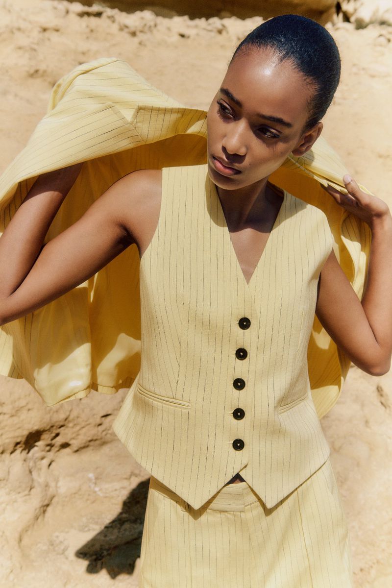 A woman in a light-yellow pinstripe vest and skirt poses in a desert landscape. The jacket is held behind her head.