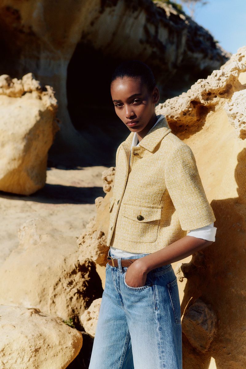 Woman in yellow jacket and blue jeans poses near sandstone rock formations in outdoor fashion shot, highlighting clothing and natural textures.