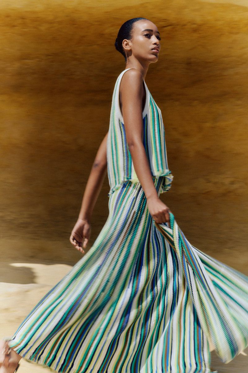 Fashion shot of a woman in a sleeveless striped top and skirt, walking in front of a yellow background with sand. Modern design.