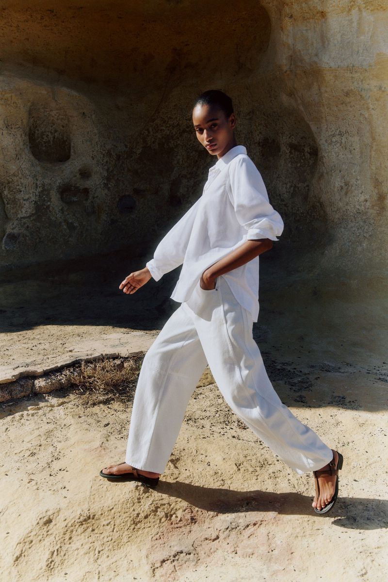 A woman walks on a sandy surface wearing a white linen shirt and wide-legged pants paired with sandals, posing against a rock formation.
