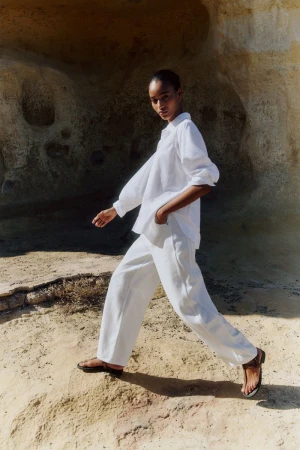 A woman walks on a sandy surface wearing a white linen shirt and wide-legged pants paired with sandals, posing against a rock formation.