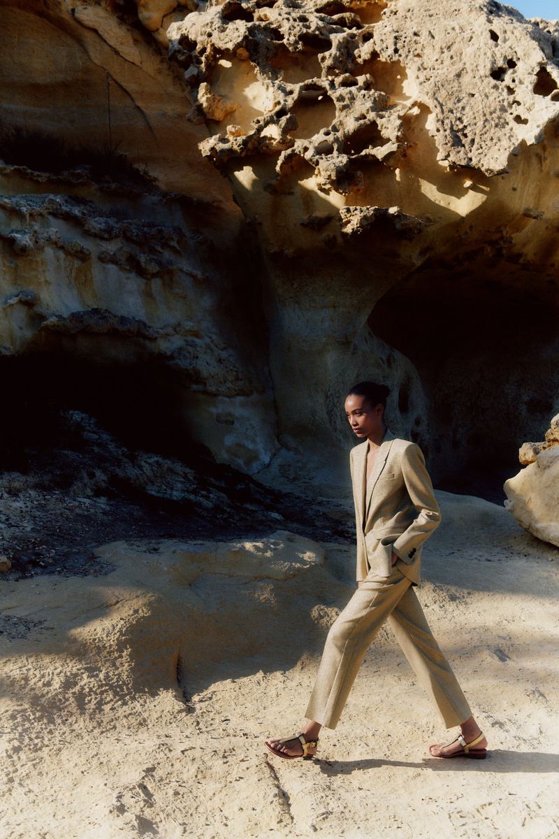 Fashion shot of a Black woman wearing a beige pantsuit walking on a beach in front of a large rock formation during the day.