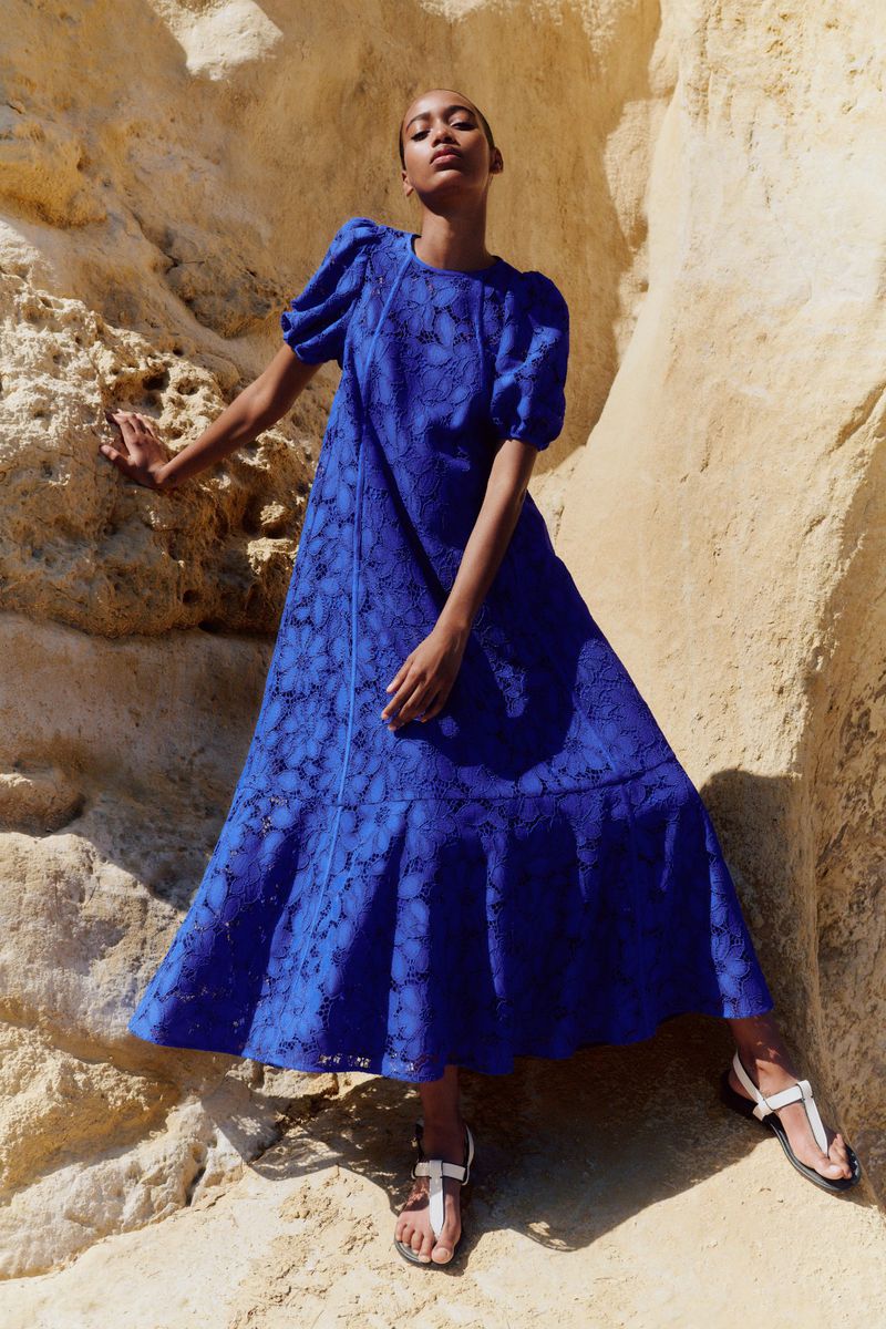 A dark-skinned woman models a flowing royal blue dress with puff sleeves and white sandals in front of a tan rock formation.