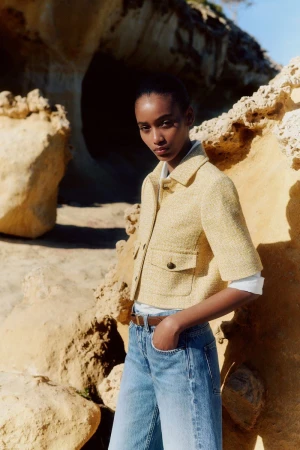 Model Manuela Sanchez wearing a cropped yellow short-sleeve jacket and blue denim jeans against sunny sandstone rocks.