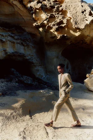 Model Manuela Sanchez wearing a tailored beige pantsuit and sandals, walking across sand in front of porous limestone cliffs.